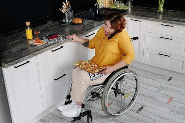 A woman in a wheelchair is talking on the phone and preparing food in a kitchen.