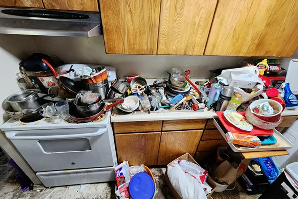 A kitchen with many dishes, pots, and pans. It is in need of cleaning and organization. There is a white stove and wooden cabinets.