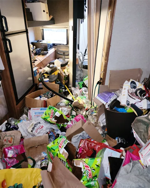 A space with a buildup of items. There are boxes and debris blocking the access and use of the space including the fridge and sink. the floor and countertop are obscured by dishes, debris, and a buildup of items.
