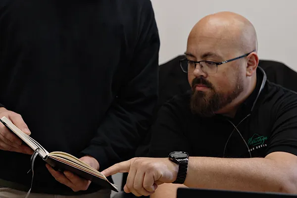 Brandon is sitting in the office pointing at something in a book that Anthony is holding.