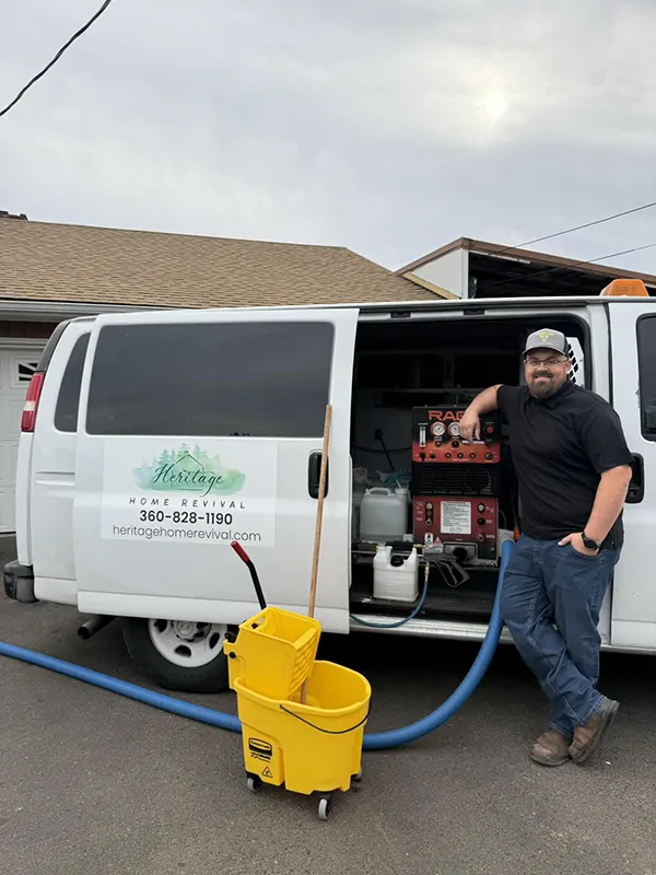 Brandon stands with the carpet cleaning van and a yellow mop bucket. The van is parked in the driveway of a house.